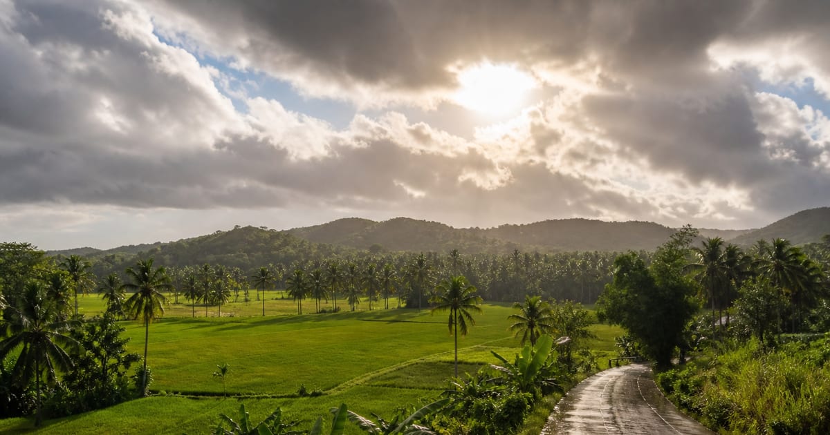 Sun breaking through rain clouds over green fields and a wet rural road in Bohol